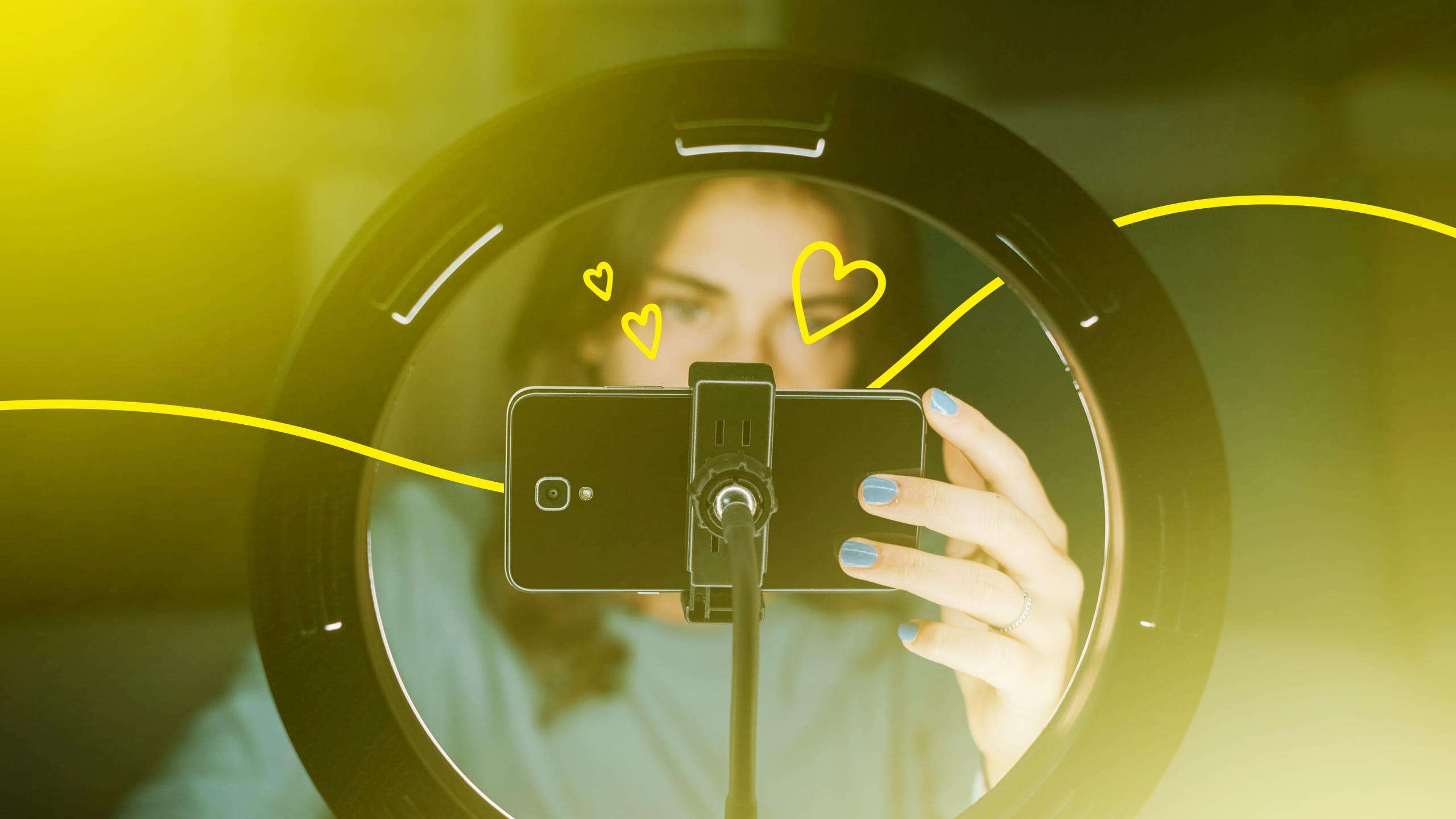 An influencer framed by a ring-light capturing a video selfie with hearts illustrated on top of the photo